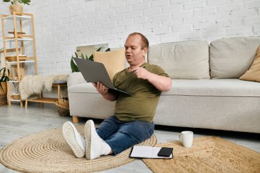 Kapsamlı bir adam oturma odasındaki halıya oturmuş, dizüstü bilgisayar kullanıyor ve nişanlı görünüyor..