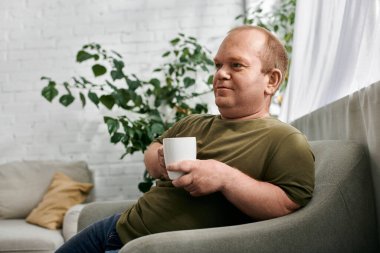 A man with inclusivity in casual attire sits on a couch, enjoying a cup of coffee in a relaxed atmosphere. clipart