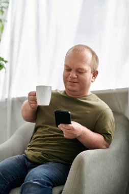 Kapsayıcı bir adam bir sandalyeye oturur, bir fincan kahvenin tadını çıkarır ve telefonunu kontrol eder..
