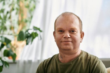 Gündelik kıyafetleri olan bir adam evinde sakin bir ifadeyle rahatça oturur..
