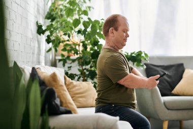 Kapsayıcı bir adam rahat bir oturma odasındaki koltukta oturur, rahat bir şekilde giyinir ve telefonunu kullanır..