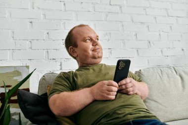 Kapsayıcılığı olan bir adam evindeki bir kanepede, elinde akıllı bir telefonla rahatlar..