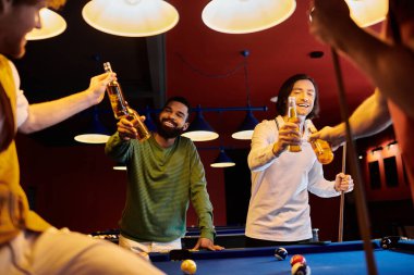 Bir grup arkadaş loş bir odada bilardo oynarken bira şişeleriyle kadeh kaldırıyor..