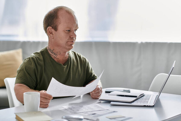 Человек с инклюзивностью сидит за своим рабочим столом, просматривая документы, в то время как ноутбук сидит поблизости.