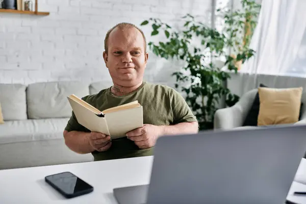 stock image A man with inclusivity in casual attire sits at a table reading a book, with a laptop and phone nearby.