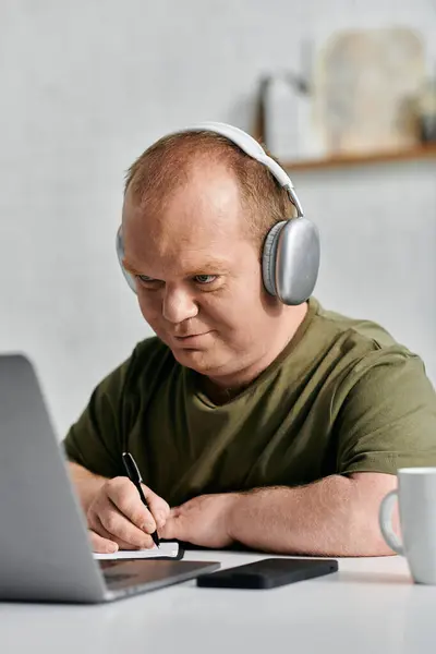 stock image A man with inclusivity in a casual shirt sits at a desk with a laptop and takes notes while wearing headphones.