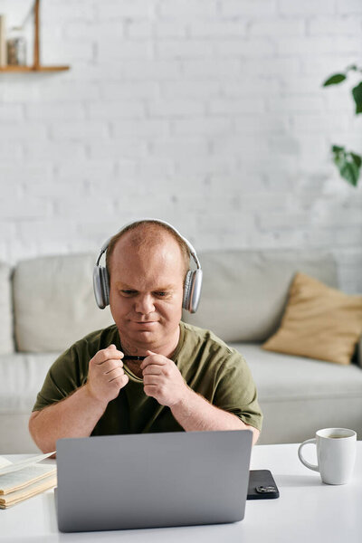 A man with inclusivity wearing headphones sits at a desk with a laptop, looking focused on his work.