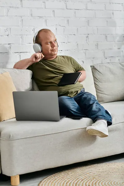 stock image A man with inclusivity in casual attire sits on a sofa at home, wearing headphones and holding a tablet.