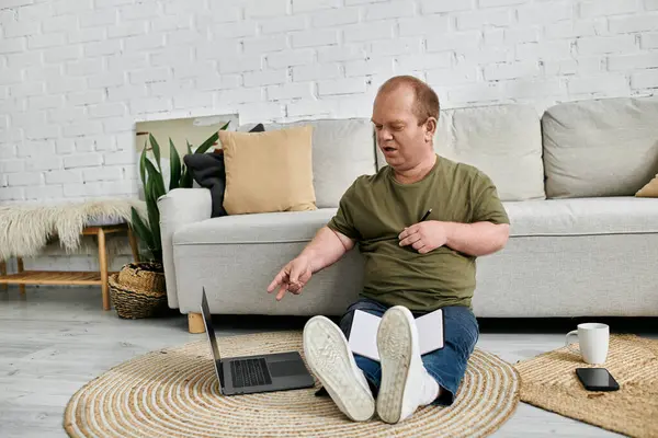 stock image A man with inclusivity sits casually on the floor in his living room, using a laptop.