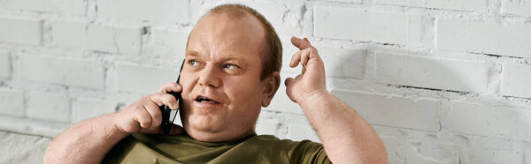 A man with inclusivity in casual attire sits in front of a white brick wall and talks on a phone.