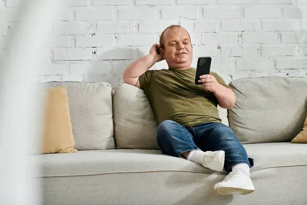 stock image A man with inclusivity relaxes on a sofa in his home, scrolling through his phone.