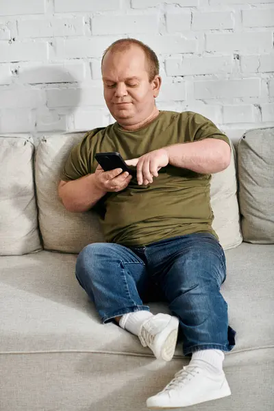 stock image A man with inclusivity sits on a couch in casual attire, looking at his phone.