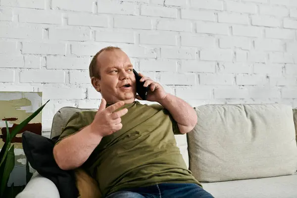 stock image A man with inclusivity in casual attire sits on a couch, talking on the phone, in a home setting.