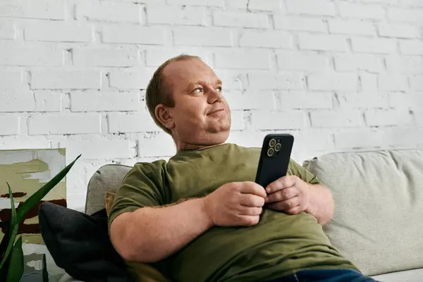 stock image A man with inclusivity relaxes on a couch in his home, holding a smartphone.
