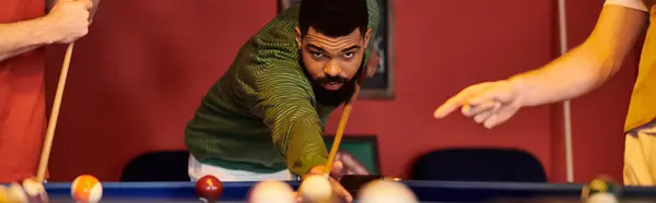 stock image A man in a green sweater lines up a shot at a billiard table while his friends watch, enjoying a casual game.