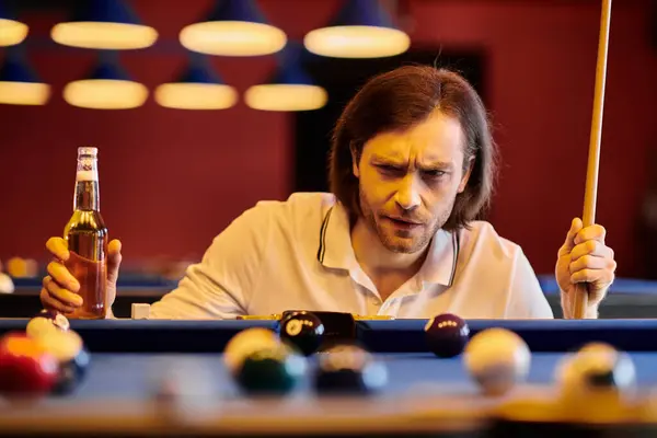 stock image A man in casual attire focuses intently on the billiard table, holding a cue stick and a beer bottle.