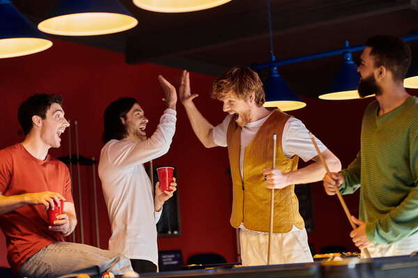 Группа друзей празднует победу за бильярдным столом, смеясь и давая пять.