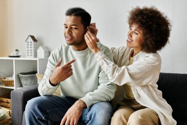 Appealing African American woman helping her husband with hearing aid while at home. clipart