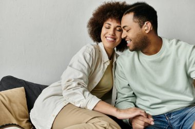 An attractive African American couple sits close together on a couch at home. clipart