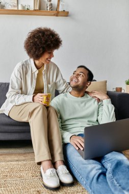 Kıvırcık saçlı Afrikalı Amerikalı kadın kocasının yanında oturuyor ve laptopuna bakıyor..