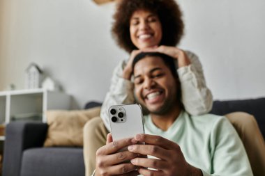 Cheerful African American woman sitting near her husband with hearing aid and looking at phone. clipart