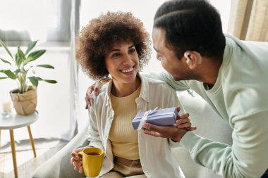 Pretty African American woman near her husband with hearing aid, gift in hands. clipart