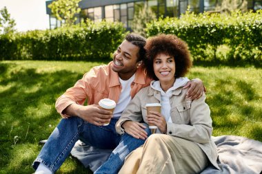 Bir Afro-Amerikalı çift parkta bir battaniyeye oturmuş, güneşin tadını çıkarıyor ve birbirlerine eşlik ediyorlar..