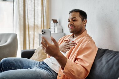 An African American man signs on his phone, while sitting on a couch in his home. clipart