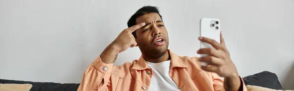 stock image An African American man sits on a couch at home, using sign language while on a video call on his phone.