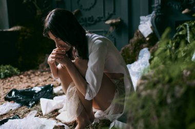 Beyaz gömlekli ve dantel etekli kadın bataklıkta oturuyor. Etrafı plastik atıklarla çevrili..