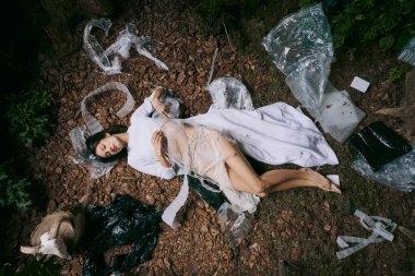 Beyaz elbiseli bir kadın ormanda yerde yatıyor, etrafı plastik çöplerle çevrili..