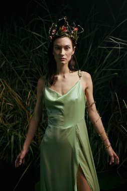 Çiçekli taçlı bir kadın bataklık bitkisinin yanında yeşil bir elbise giyiyor..