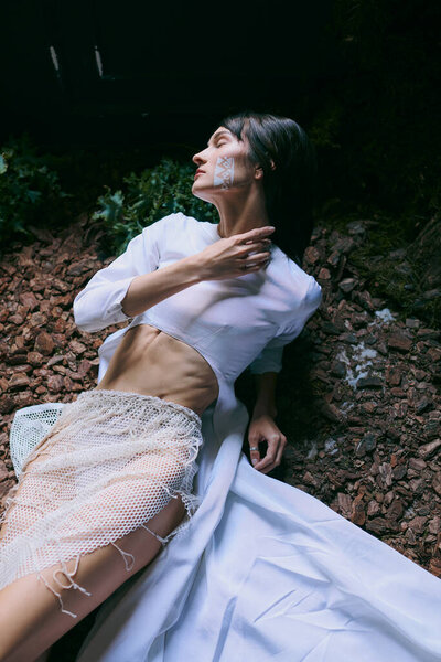 A woman in a white dress poses on the ground near a swamp.