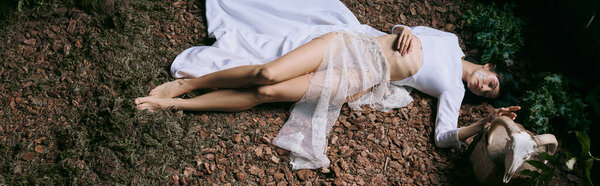 A woman in white clothing lies on ground, surrounded by foliage.