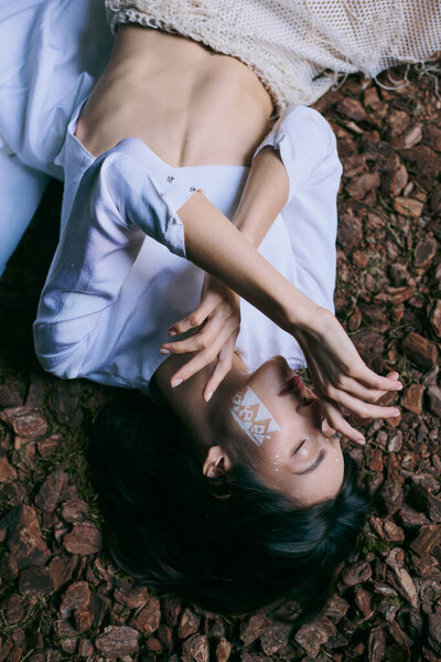 A woman in white lies near swamp, her face obscured by a geometric design.