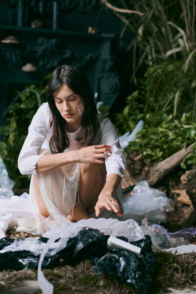 A woman in a white dress poses amidst a swamp filled with plastic garbage.