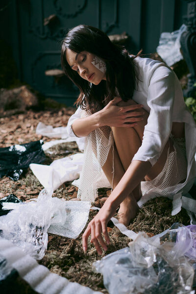 Женщина в белом платье позирует посреди пластикового мусора в болотистой местности.