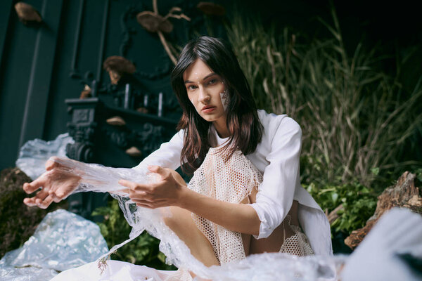 A woman in a white dress poses amidst a swamp filled with plastic waste.