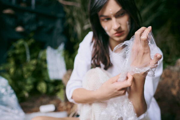 A woman in a white dress poses with plastic wrap in a swamp filled with plastic garbage.