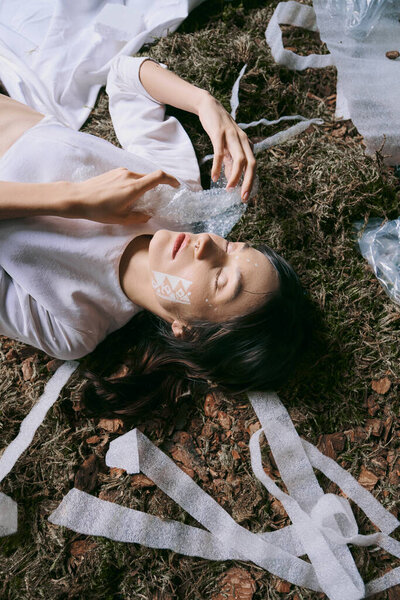 A woman lays surrounded by plastic waste, her eyes closed and expression solemn.