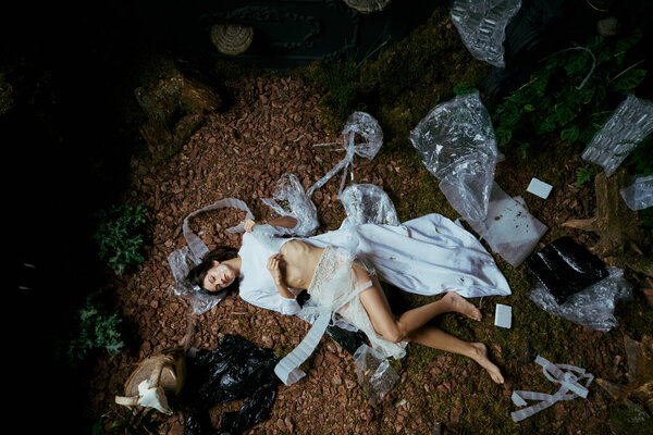 Женщина в белой одежде лежит посреди болота, задушенного пластиковым мусором.