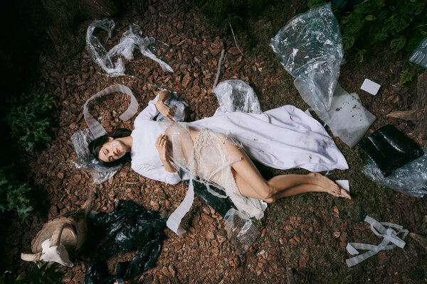 Женщина в белом платье лежит на лесной подстилке, окруженная пластиковым мусором.