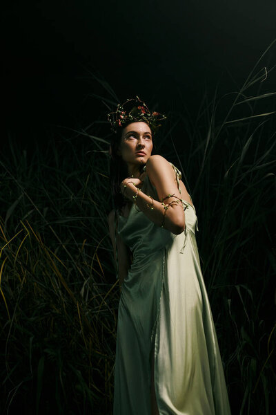 A woman in a flowing green dress poses near a swamp at night.