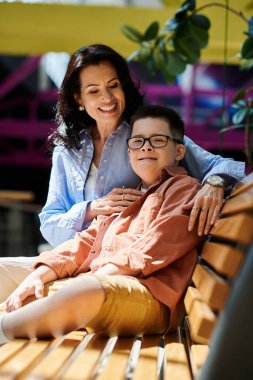 A mother and her son with Down syndrome share a heartwarming moment on a bench in a shopping mall. clipart