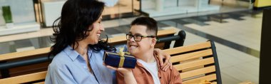 A mother and her son with Down syndrome share a heartwarming moment while sitting on a bench in a shopping mall. clipart