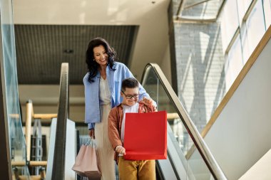 A mother and her son with Down syndrome enjoy a day of shopping at the mall, riding the escalator and carrying shopping bags. clipart