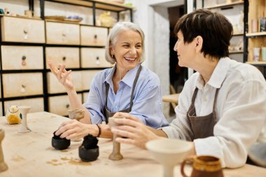 Çömlek atölyesinde iki yetişkin kadın, kil projeleri üzerinde çalışıyor ve bir kahkahayı paylaşıyorlar..