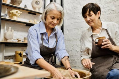 Two mature women in a pottery studio, one working on the wheel while the other observes. clipart