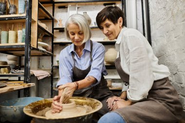 İki kadın çömlek atölyesinde el işi yapıyor, bir kadın çömlek tekeri üzerinde çalışıyor..
