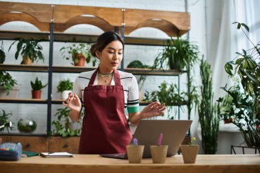 A beautiful Asian woman, the owner of a small plant shop, stands behind a wooden counter and uses a laptop in her plant-filled shop. clipart